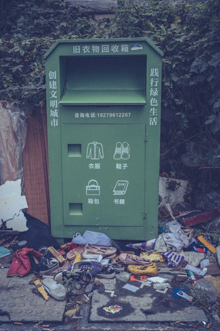 A green, weathered old clothing collection box positioned outdoors against a backdrop of trees and foliage, with visible trash scattered around its base. The box has Chinese characters and a contact p