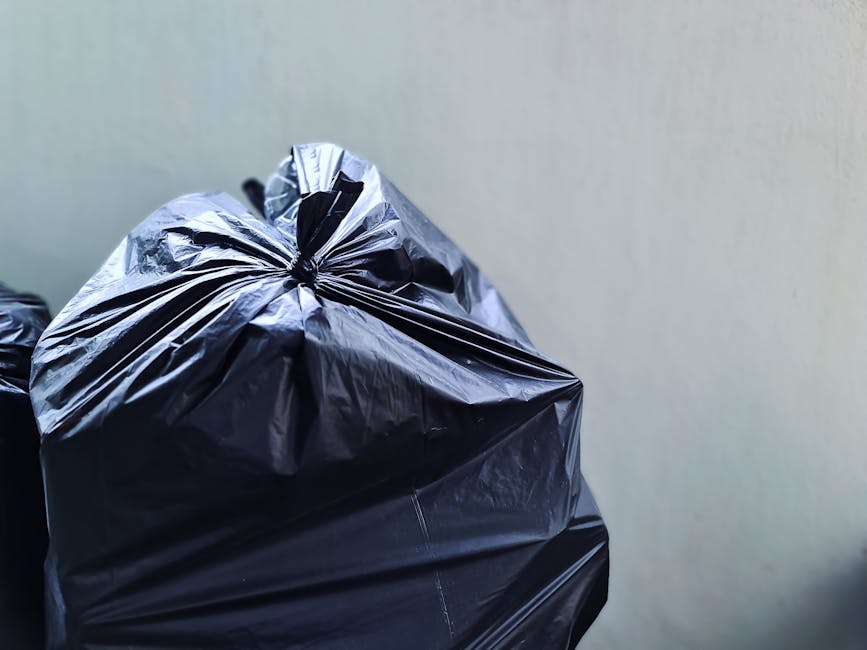 A middle-aged man with dark hair, wearing a black T-shirt with graphic text, is bent over placing a large white plastic bag filled with waste into a metal rubbish bin on a paved sidewalk. The bin, mad